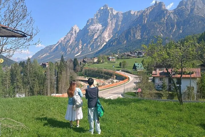 Two travelers on a green meadow looking up at a jagged dolomite peak above a valley town